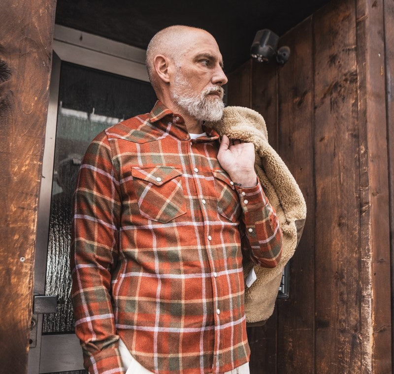 Man in a plaid flannel western shirt with a shearling-collar jacket over his shoulder, in front of a wooden door. Photo @chesapeakesofficial
