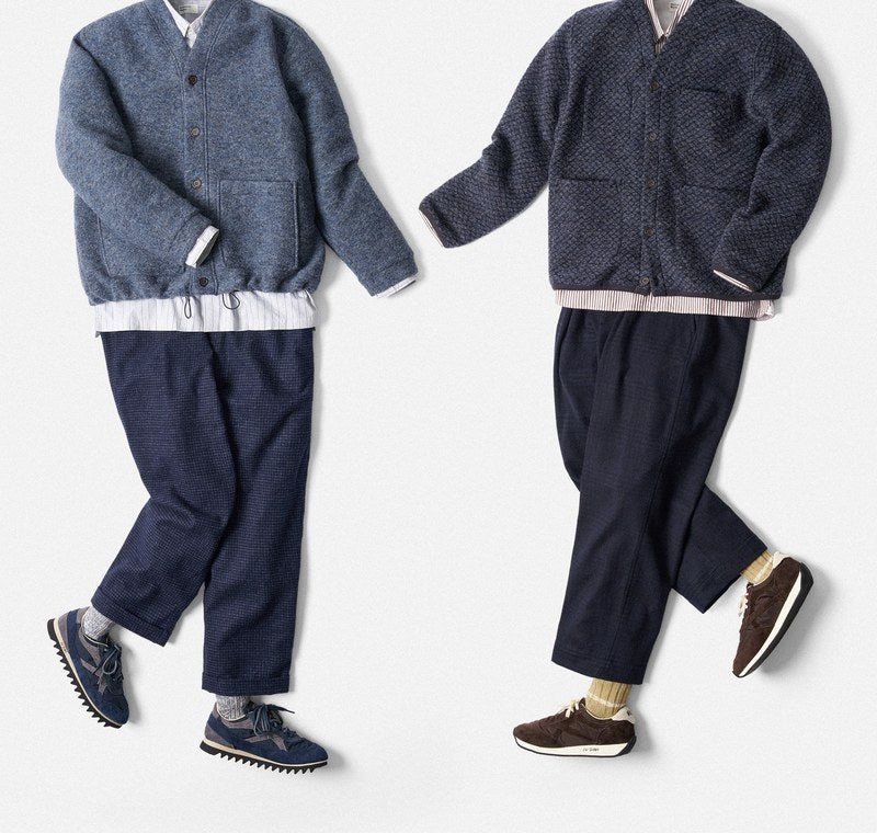Two Universal Works outfits laid flat: dark and light blue wool cardigans, white shirt, blue trousers and sneakers.